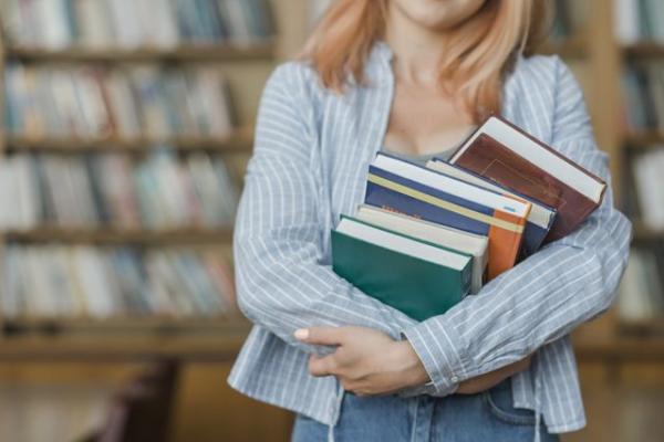 Женщина держит стопку книг в библиотеке.