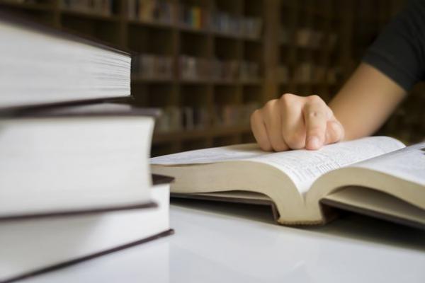 Человек читает открытую книгу за столом рядом со стопкой книг.